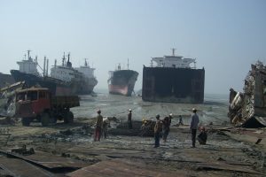 ship breaking india with workers and truck on the side, scrap materials strewn across with a few large ships settled on the beach