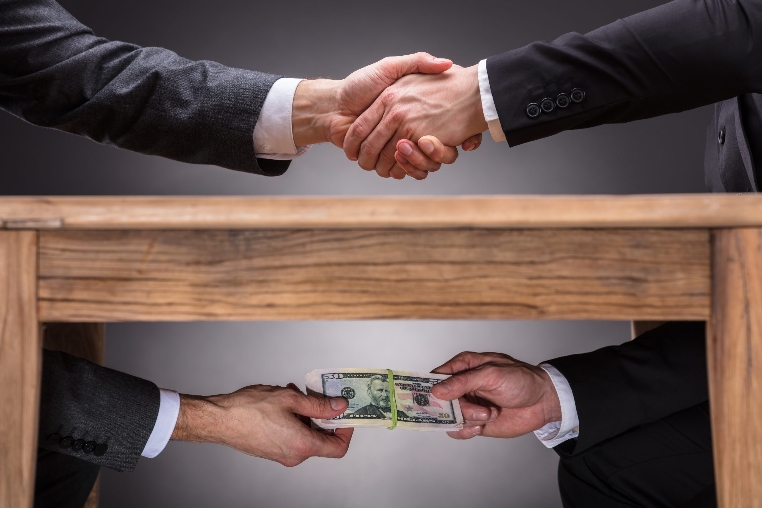 Close-up Of Two Businesspeople Shaking Hand And Taking Bribe Under Wooden Table On Grey Background