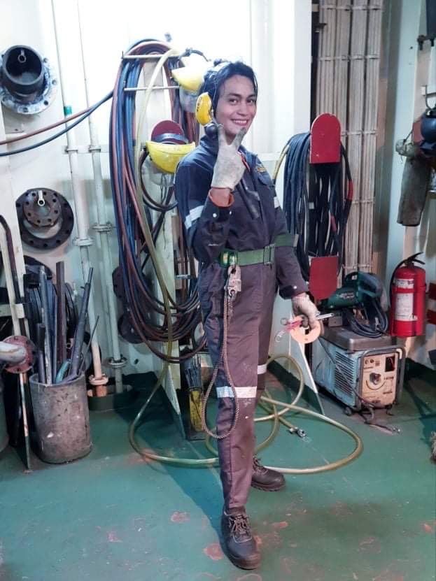 Dyosa in uniform, striking a pose inside the ship, with hanging tubes and pipes in the background