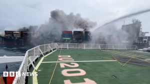 A ship is on fire. Thick dark smoke rises from the containers on its deck. In the foreground, we see the deck of another vessel with railings and a large green area marked “0205.” A strong stream of water sprays from the boat, aiming at the fire.